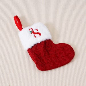 Può includere: Una piccola calza di Natale rossa con un polsino bianco e soffice. La calza ha un design a trecce e un anello rosso per appenderla. Il polsino presenta una "S" ricamata con un bastoncino di zucchero e agrifoglio.