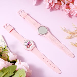 May include: Two light pink wristwatches with silicone bands. One watch shows the front face with a white dial featuring a colorful cartoon character graphic and black numerals. The other watch is flipped to show the smooth, circular silver metal case back.