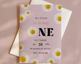 以下が含まれることがあります： ピンク色の招待状。デイジーのイラストと「BLOOM is turning ONE」の文字が特徴です。10月開催のイベントの日時と場所、RSVPの電話番号が記載されています。白い封筒に入っています。