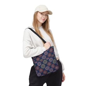 Könnte beinhalten: Eine marineblaue Tragetasche mit einem sich wiederholenden Muster aus kreisförmigen, sternartigen Designs in Rot-, Weiß- und Goldtönen. Die Tasche hat schwarze Schulterriemen und wird über der Schulter getragen, wobei die detaillierten Blumenmotive sichtbar sind.