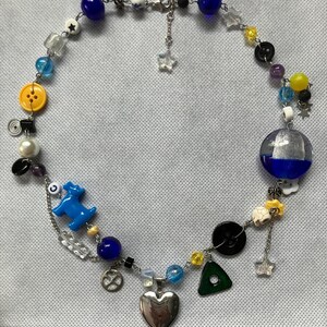 May include: A handmade necklace featuring a variety of beads in blue, yellow, clear, and black. The necklace includes a silver heart pendant, a blue dog charm, and a yellow button. The necklace is approximately 18 inches long.