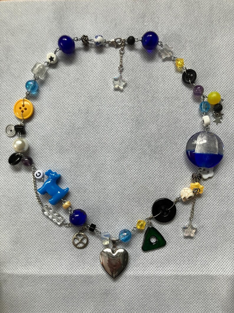 May include: A handmade necklace featuring a variety of beads in blue, yellow, clear, and black. The necklace includes a silver heart pendant, a blue dog charm, and a yellow button. The necklace is approximately 18 inches long.