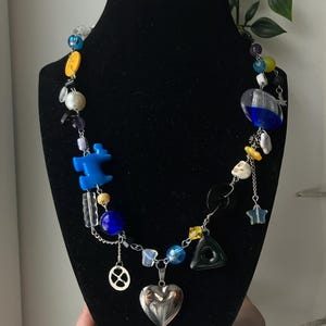 May include: A colorful necklace with various beads and charms, including blue, yellow, and clear beads, a silver heart pendant, and a star charm. The necklace is displayed on a black mannequin.