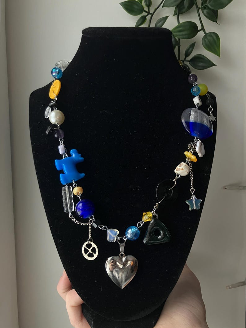 May include: A colorful necklace with various beads and charms, including blue, yellow, and clear beads, a silver heart pendant, and a star charm. The necklace is displayed on a black mannequin.
