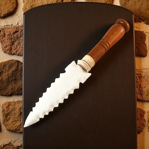May include: A decorative knife with a white, jagged blade and a wooden handle. The handle is brown with a rounded end and is attached to the blade with a wrapped, beige material. The knife is displayed against a dark brown background.