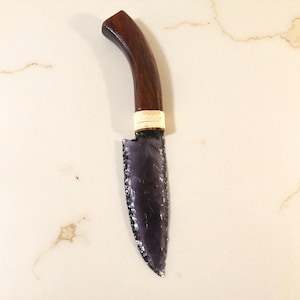 May include: A handcrafted knife featuring a dark purple blade and a curved, dark wood handle. The blade has a decorative edge and is connected to the handle with a white band. The knife is displayed on a light-coloured surface.
