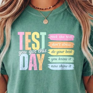 Può includere: Una maglietta color verde acqua con le parole "TEST DAY" in grandi lettere colorate. La maglietta presenta anche le frasi "you got this", "rock the test", "don't stress", "do your best", "you know it" e "now show it".