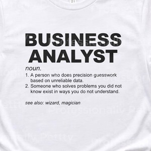 Op de afbeelding: Wit t-shirt met de tekst "BUSINESS ANALYST" in dikke zwarte letters. Daaronder staat een definitie van de term. Onderdeel van een kledingcollectie.