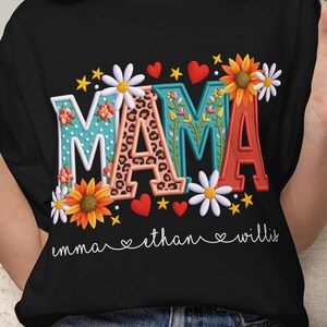 May include: Black t-shirt with the word "MAMA" in colorful floral and leopard print letters. The shirt also features the names "emma", "ethan", and "willis" below the word "MAMA".