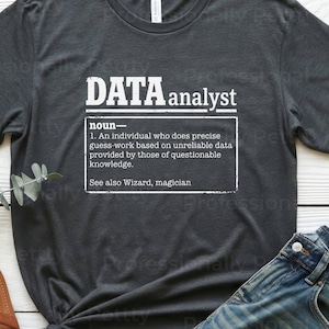May include: Dark grey t-shirt featuring the text "DATA analyst" and a definition. The shirt is styled with blue jeans and brown sandals. The text on the shirt defines a data analyst.