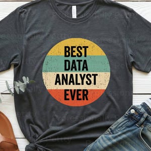 May include: Dark heather gray t-shirt with a circular graphic that reads "BEST DATA ANALYST EVER" in white text. The graphic has yellow, teal, and orange horizontal stripes. The shirt is paired with blue jeans and brown sandals.