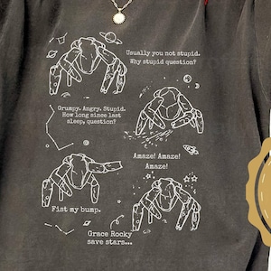 May include: A charcoal gray short-sleeve t-shirt featuring white line art illustrations of four crab-like creatures. The graphic includes text snippets: Usually you not stupid, Why stupid question, Grumpy, Angry, Stupid, Fist my bump, Amaze! Amaze! Amaze!, and Grace Rocky save stars.