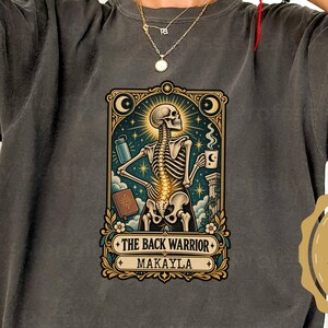 Puede incluir: Una camiseta de manga corta gris carbón con un gráfico central de un esqueleto en un marco de carta de tarot. El esqueleto sostiene una taza y una botella, con el texto THE BACK WARRIOR y MAKAYLA impreso debajo.