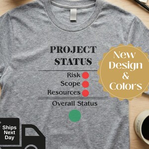 May include: A heather gray t-shirt with the words "PROJECT STATUS" and status indicators. The shirt also has a "New Design & Colors" sticker and a "Ships Next Day" graphic.