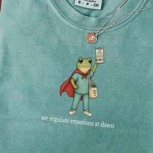 Op de afbeelding: Een teal Comfort Colors T-shirt met een cartoon kikker superheld die een klembord en een tas vasthoudt. De tekst "we regulate emotions at dawn" staat onder de kikker gedrukt. Een zilveren ketting met een hanger ligt op het T-shirt.