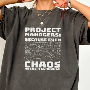 May include: Dark gray t-shirt with white text that reads "PROJECT MANAGERS: BECAUSE EVEN CHAOS NEEDS A SCHEDULE." The shirt features a graphic of mathematical symbols and equations. The shirt has short sleeves and a relaxed fit.