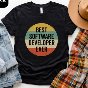 Może przedstawiać: Czarny t-shirt z napisem "BEST SOFTWARE DEVELOPER EVER" w stylu retro. Tekst znajduje się w okręgu z żółtymi, zielonymi, niebieskimi i czerwonymi paskami. Koszulka jest prezentowana z kurtką dżinsową i koszulą w kratę.