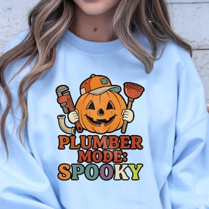 May include: A light blue crewneck sweatshirt featuring a graphic of a smiling orange jack-o'-lantern wearing a baseball cap and holding a pipe wrench and a plunger. The text below the pumpkin reads "PLUMBER MODE: SPOOKY" in colorful letters.