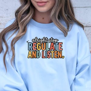 Op de afbeelding: Een lichtblauw sweatshirt met ronde hals en de tekst "alright stop REGULATE AND LISTEN" in een meerkleurig, retro lettertype. De afbeelding bevat kleine ster- en hartaccenten en is gedrukt op het midden van de borst van het kledingstuk.
