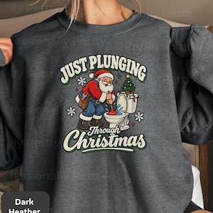 May include: A dark heather gray crewneck sweatshirt featuring a graphic of Santa Claus using a plunger on a toilet. The text reads JUST PLUNGING THROUGH CHRISTMAS. The design includes a small Christmas tree on the toilet tank and white snowflakes.