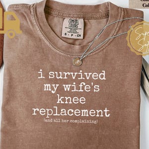 May include: A brown short-sleeved t-shirt with white text that reads: I SURVIVED my wife's knee replacement (and all her complaining). The shirt features a Comfort Colors brand label at the inner neckline and a silver-toned necklace resting on top.