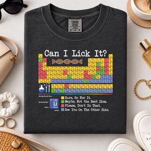 Puede incluir: Una camiseta de cuello redondo gris carbón con un gráfico colorido de la tabla periódica y el texto Can I Lick It? encima. El diseño incluye una cadena de ADN, un vaso de precipitados y una leyenda que explica los niveles de seguridad.