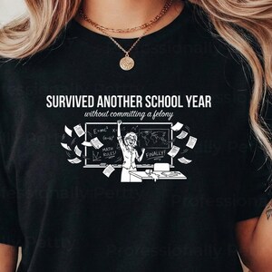 May include: Black crew neck t-shirt featuring the text "SURVIVED ANOTHER SCHOOL YEAR without committing a felony" and a cartoon illustration of a person celebrating in front of a chalkboard with flying papers.