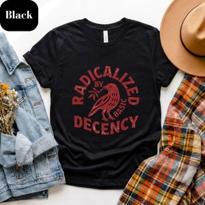 Könnte beinhalten: Schwarzes T-Shirt mit roter Grafik, die einen Vogel mit einem Zweig zeigt, umgeben von den Worten "RADICALIZED BY BASIC DECENCY". Das Shirt wird mit einer Jeansjacke, einem karierten Hemd und einem braunen Hut präsentiert.