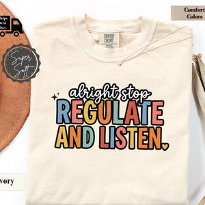 Op de afbeelding: Een ivoorkleurig t-shirt met de tekst "alright stop REGULATE AND LISTEN." in een meerkleurig, retro-geïnspireerd lettertype. Het shirt heeft een ronde hals, een Comfort Colors merklabel in de kraag en een klein hartvormig icoontje.