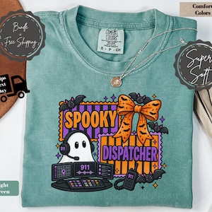 Könnte beinhalten: Ein hellgrünes T-Shirt mit einer Grafik eines Geistes, der ein Headset an einer Computerkonsole trägt, mit dem Text Spooky Dispatcher. Das Design enthält eine orangefarbene Schleife mit Fledermausmustern sowie violette und orangefarbene Streifen.