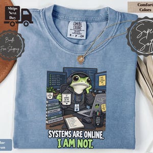 May include: A blue jean colored t-shirt featuring a graphic of a frog wearing headphones and a hoodie, sitting at a desk with computer monitors, a laptop, and a mug that reads IT WORKS ON MY MACHINE. The text reads SYSTEMS ARE ONLINE. I AM NOT.