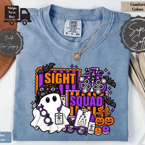 May include: A blue jean coloured t-shirt featuring a graphic print of a ghost wearing a bandana and glasses, surrounded by bats, pumpkins, an eye chart, and an optometry phoropter. The text SIGHT SQUAD is printed in bold purple and orange letters.