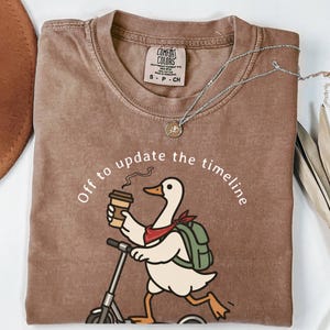 May include: Brown Comfort Colors t-shirt with a graphic of a duck riding a scooter, holding coffee, and wearing a backpack. Text reads "Off to update the timeline." A silver necklace and a brown hat are also visible.