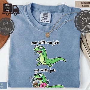 May include: A blue jean coloured short-sleeve t-shirt featuring a graphic of a green lizard. The top graphic shows the lizard crying with the text me with no job. The bottom graphic shows the lizard crying while holding a coffee and a donut with the text me with job. The shirt has a crew neckline and a Comfort Colors brand label.