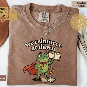Könnte beinhalten: Ein espressofarbenes T-Shirt mit einer Grafik eines grünen Frosches, der einen roten Umhang trägt. Der Frosch hält ein Schild mit der Aufschrift "FIRST THEN" und einen gelben Stern. Der Text "we reinforce at dawn" steht darüber, ergänzt durch Sterne und Bücher.