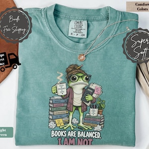 Può includere: Una t-shirt a maniche corte verde chiaro con la grafica di una rana con occhiali e cardigan, che tiene in mano una tazza di caffè con l'etichetta Fueled by Spreadsheets & Coffee e una calcolatrice, con il testo Books Are Balanced. I Am Not.