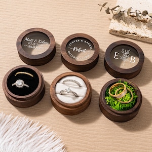 May include: Six wooden ring boxes with clear lids. Each box contains a ring, with some boxes displaying text such as names and dates. The rings are gold and silver, and the boxes are lined with black or white fabric and green moss.