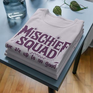 May include: A light pink t-shirt featuring dark purple text that reads MISCHIEF SQUAD and we are up to no good. The design includes small starburst accents, a series of small footprints, and a feather graphic printed on the front of the garment.