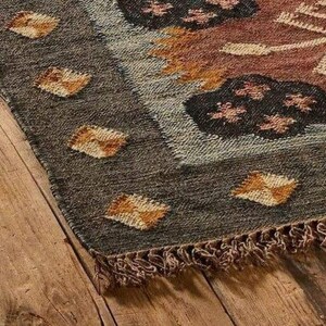 May include: Close-up of a woven rug with a geometric pattern. The rug features a dark gray border with diamond-shaped accents in shades of brown and gold. The central design includes floral and abstract shapes in brown, orange, and white.