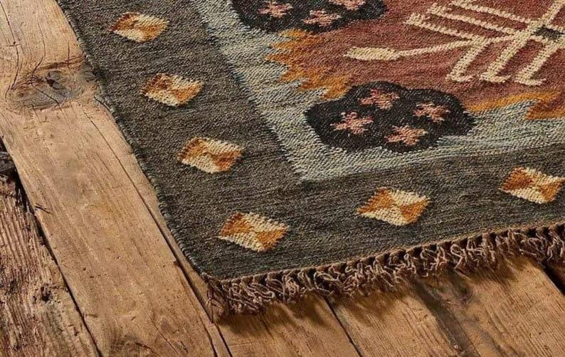 May include: Close-up of a woven rug with a geometric pattern. The rug features a dark gray border with diamond-shaped accents in shades of brown and gold. The central design includes floral and abstract shapes in brown, orange, and white.