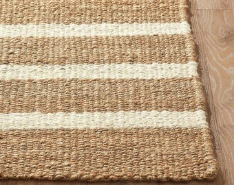 May include: A rectangular rug with a natural, woven texture. The rug features alternating horizontal stripes of tan and off-white. The rug is made of a durable material and is suitable for indoor use. The rug is placed on a wooden floor.