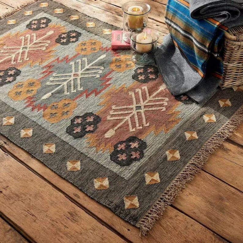 May include: A rectangular area rug with a geometric pattern in shades of gray, orange, pink, and cream. The rug has a fringed edge and is placed on a wooden surface. Decorative candles and a basket with a blanket are in the background.