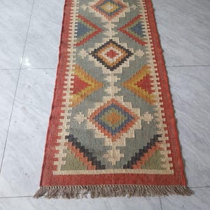 Puede incluir: Una alfombra de pasillo rectangular tejida con un patrón geométrico. La alfombra presenta un diseño central en tonos de azul, amarillo y rojo, enmarcado por un borde rojo y flecos. Sus dimensiones son de aproximadamente 60 x 150 cm.