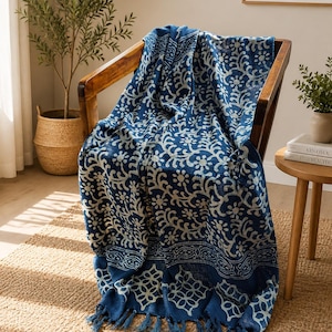 May include: A deep blue throw blanket featuring a white floral and geometric block print pattern. The fabric has a textured appearance and is finished with knotted fringe tassels along the bottom edge, draped over a wooden chair.