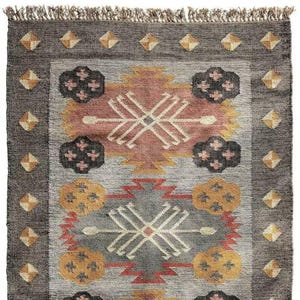 May include: A rectangular rug with a geometric pattern in shades of gray, peach, yellow, and black. The rug has a dark gray border with diamond-shaped accents and a fringed edge. The central design features stylized shapes and cross-like patterns.