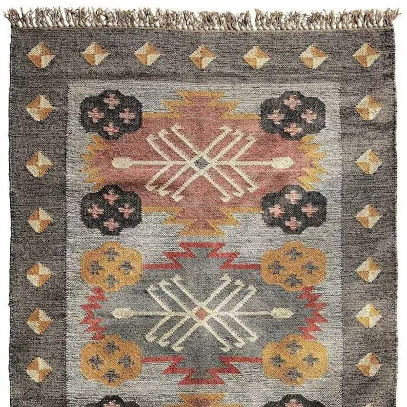 May include: A rectangular rug with a geometric pattern in shades of gray, peach, yellow, and black. The rug has a dark gray border with diamond-shaped accents and a fringed edge. The central design features stylized shapes and cross-like patterns.