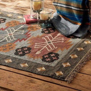 May include: A rectangular, patterned rug with a fringed edge. The rug features a geometric design in shades of gray, brown, orange, and red. Two glass candle holders with lit candles and a folded blanket are visible.