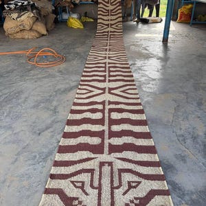 May include: A long, narrow runner rug featuring a repeating geometric pattern in dark brown and cream. The design includes symmetrical, angular shapes and lines running the length of the rug, finished with a fringed edge at the bottom end.