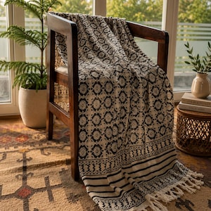 Peut inclure: Un jeté de lit noir et crème orné d'un motif floral répétitif, de bordures à rayures horizontales et de franges nouées. Le textile est drapé sur une chaise en bois foncé, mettant en valeur un design imprimé géométrique et botanique.