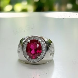 Op de afbeelding: Een zilveren ring met een grote, ovaal geslepen, helderrode edelsteen als middelpunt. De edelsteen is omgeven door een halo van kleinere, heldere stenen. De ring heeft een brede band met stervormige patronen aan de zijkanten.
